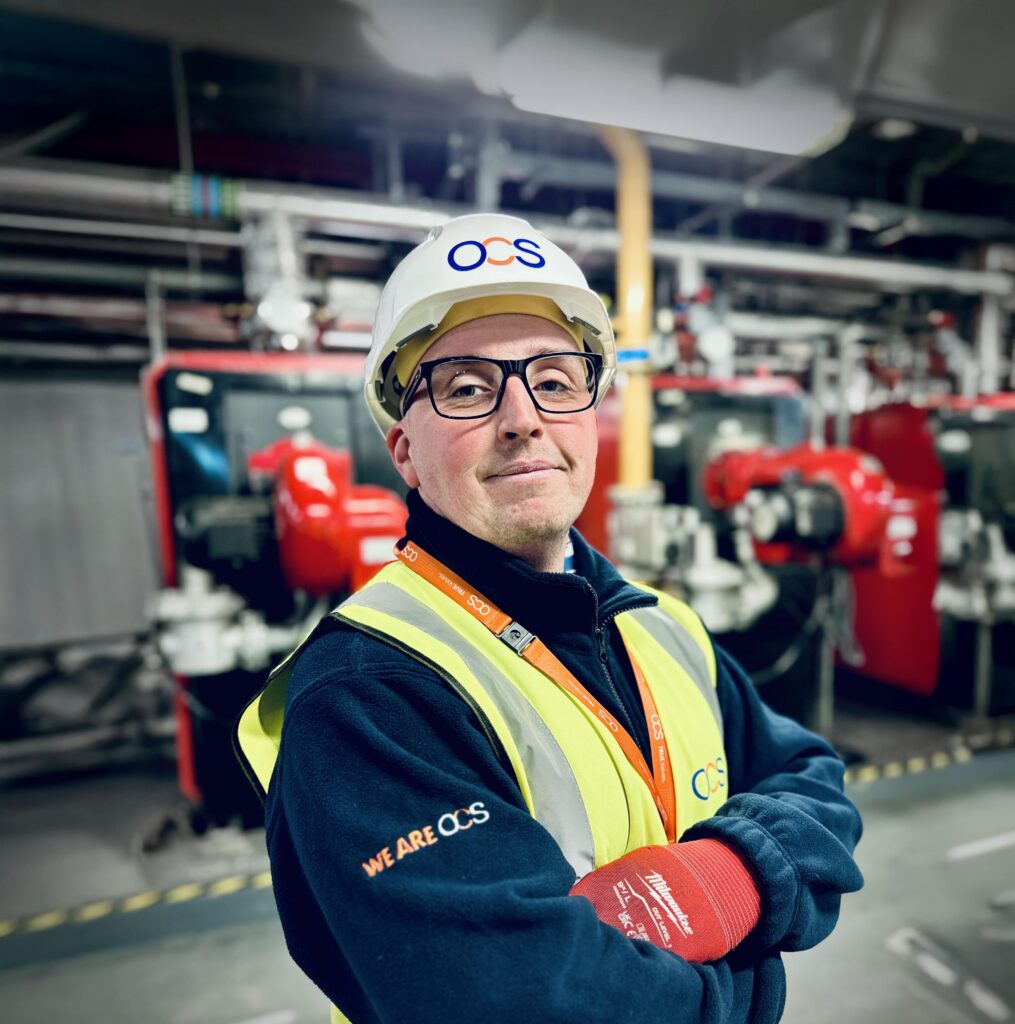 A person wearing a hard hat and safety vest stands confidently with crossed arms in an industrial setting. Machinery and pipes are visible in the background. The person is wearing glasses and appears to be in a professional work environment.